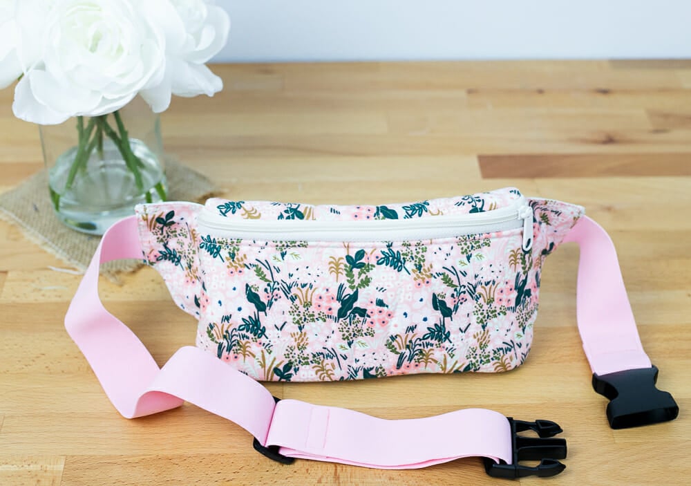 A classic, crescent-shaped waist bag featuring a front zipper closure and an adjustable black webbing strap, shown in a vibrant, multi-colored geometric pattern.