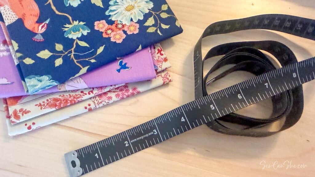 A black flexible measuring tape coiled on a wooden table with colorful fabric scraps in the background.