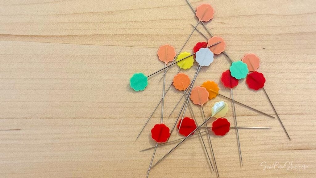 A variety of colorful straight pins with flat and round heads scattered on a wooden surface.