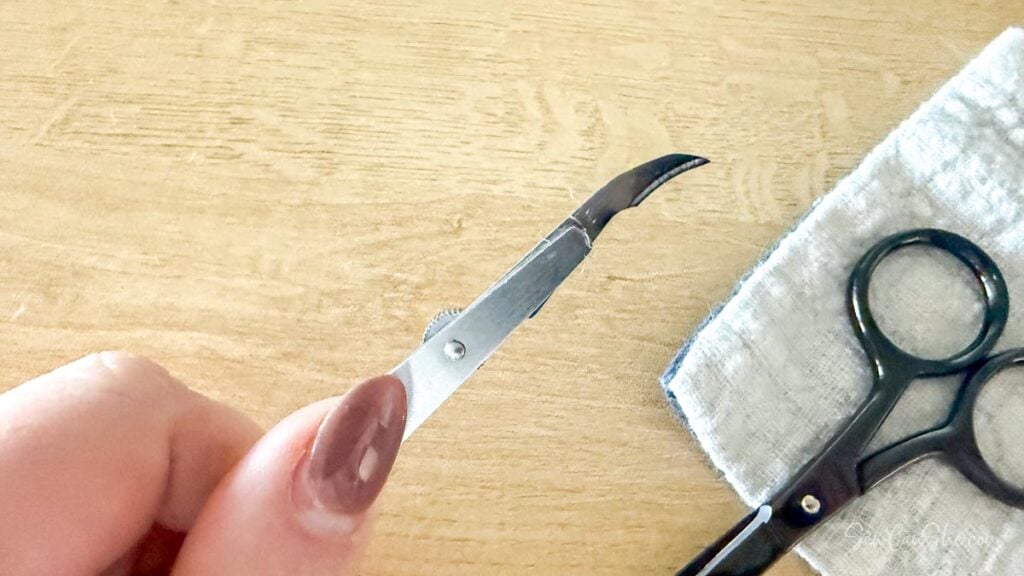 A hand holding a silver surgical-grade seam ripper to carefully remove stitches from fabric.