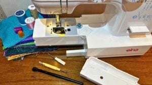 Overhead view of a sewing machine workspace with a screw driver, lint brushes, thread, and a new needle. The bobbin case is exposed and ready for a dusting; representing a regular machine maintenance routine.
