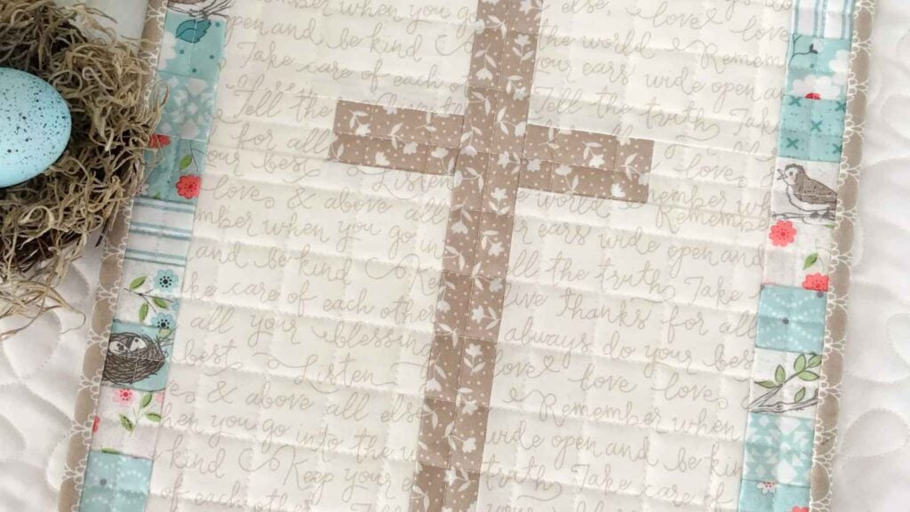 A religious Easter mini quilt titled "He Is Risen" by Carried Away Quilting. The design features a simple fabric cross centered within a scrappy pastel border, perfect for a spring wall hanging or handmade Easter decor