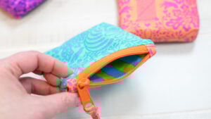A close-up of a hand holding a handmade bright blue fabric zipper pouch with a contrasting orange zipper, showcasing the professional finish and interior lining of a free sewing pattern.